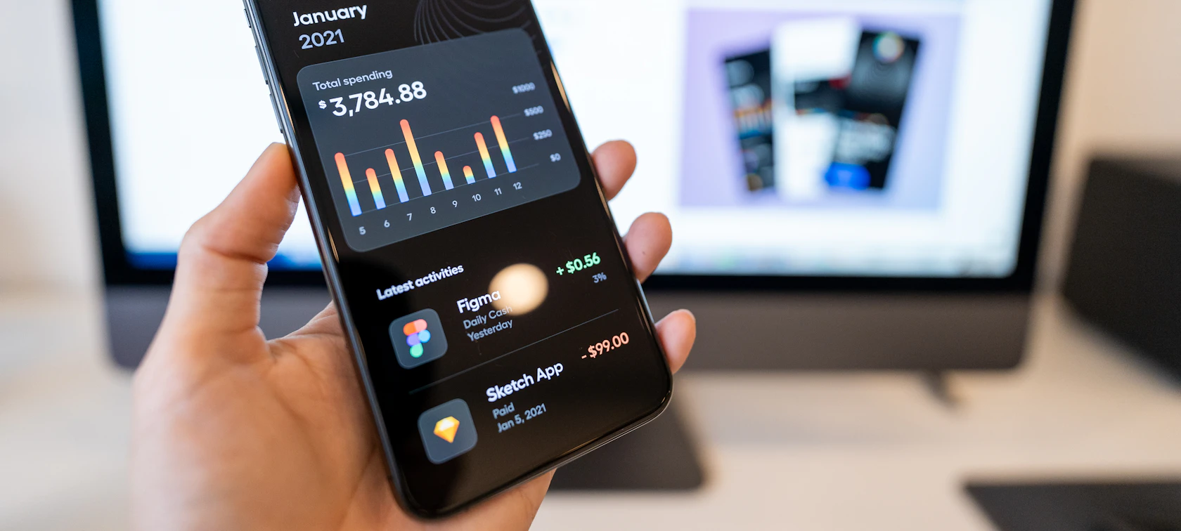 Hand holding a smartphone showing a mobile app interface in front of a design monitor