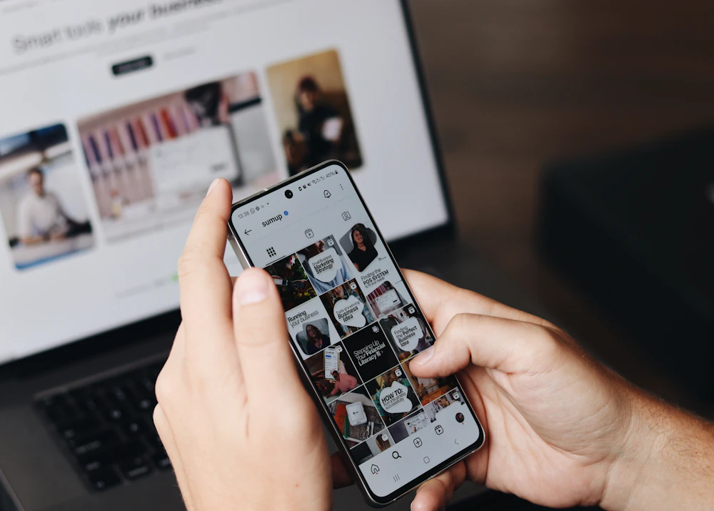 Person scrolling a social media feed on a phone in front of a laptop